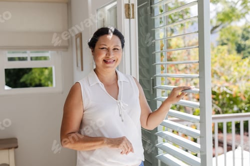 Preview: Smiling woman enjoying sunlight at home, standing by open window shutters