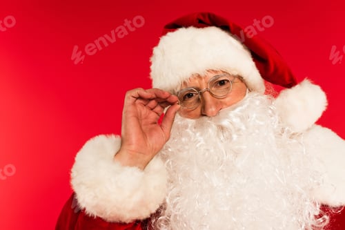 Preview: Portrait of bearded santa claus touching eyeglasses isolated on red