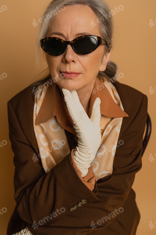 Preview: senior woman in sunglasses and white glove looking at camera isolated on beige