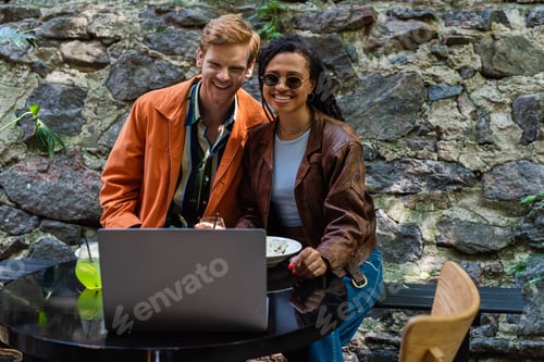 Preview: cheerful interracial friends watching movie while having lunch in cafe on terrace