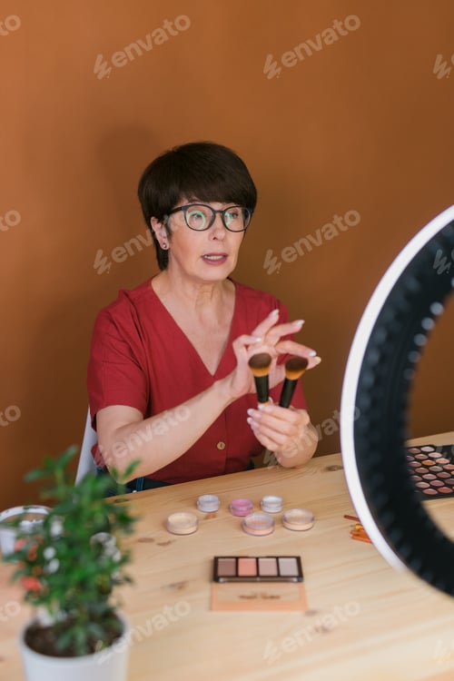 Preview: Middle-aged female beauty blogger or influencer with makeup product recording video in room at home