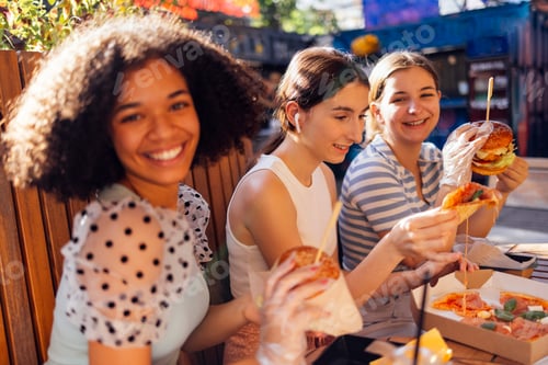 Preview: Cute smiling teenage girls are sitting in open air cafe and eating fast food.