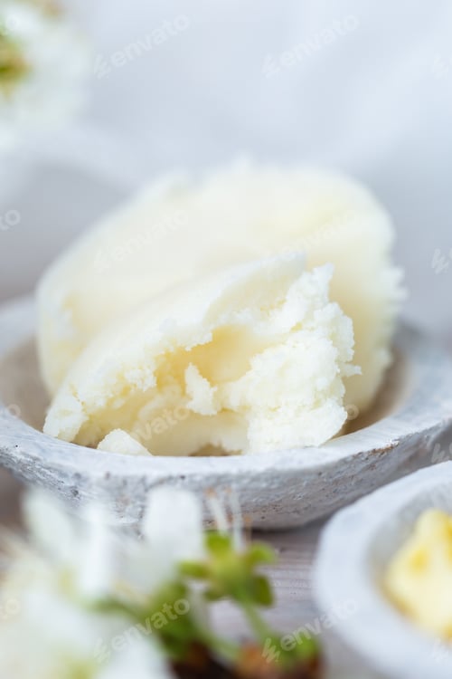 Preview: Vertical selective focus shot of shea butter in a saucer and some white flowers on a white table top