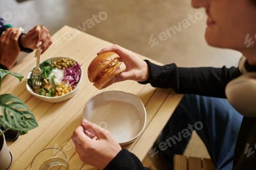 Preview: cropped young man enjoying vegan meal while holding burger with tofu near blurred woman eating salad