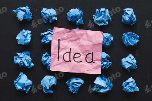 Preview: top view of 'idea' word written on sticky note with blue crumpled paper balls on black background,