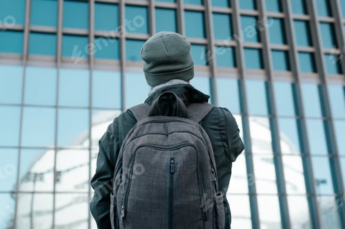 Preview: Unrecognizable man in outerwear with backpack standing on background of glass wall,