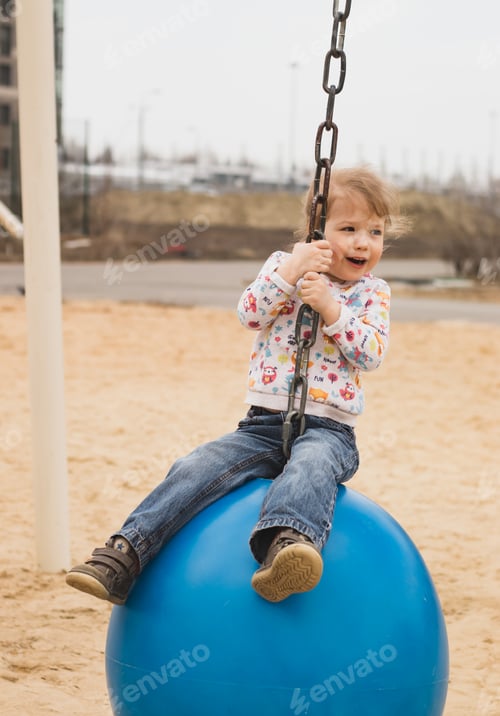 Vista previa: A little cheerful girl having fun, riding on a blue swing ball in a city park