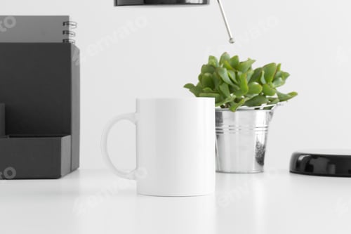 Preview: White mug mockup with a lamp, succulent plant and workspace accessories on a white table.