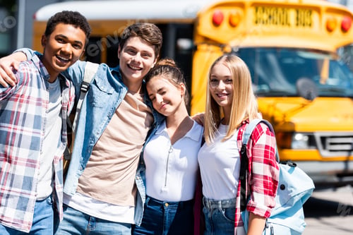 Preview: group of teen happy scholars looking at camera and embracing while standing in front of school bus