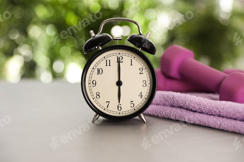 Preview: Alarm clock, towel and dumbbells on grey table against blurred background. Morning exercise