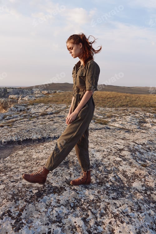 Preview: Woman in green jumpsuit standing on rock in field, traveling in nature beauty and adventure concept