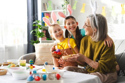 Preview: Happy family, smiling grandmother and two little girls painting easter eggs wearing bunny ears
