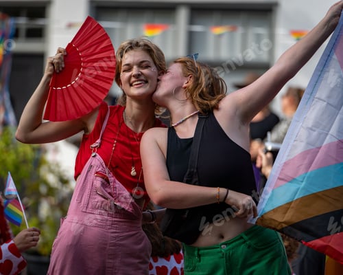 Preview: Young girl holding a flag and kissing a girl on her cheek with smiles during the canal pride 2024