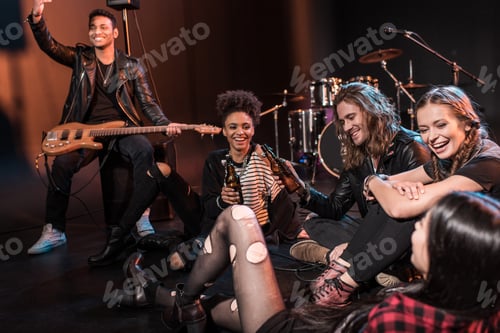 Preview: Young rock and roll band sitting together and drinking beer after concert on stage