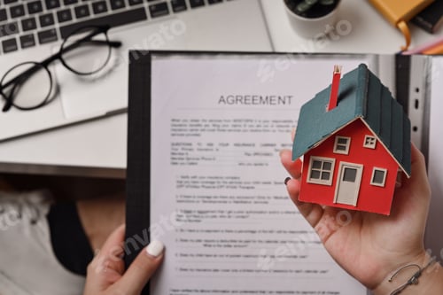 Preview: Woman holding house model and agreement contract in hand.