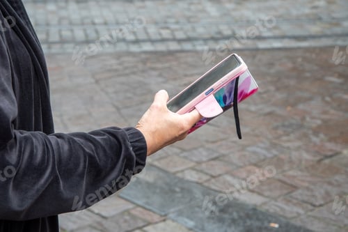 Preview: unrecognizable woman in black blouse uses smartphone in pink case on the street in the city
