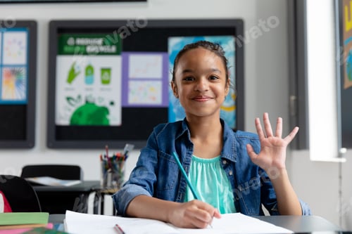 Preview: Portrait of happy biracial schoolgirl sitting at desk in class, waving, with copy space