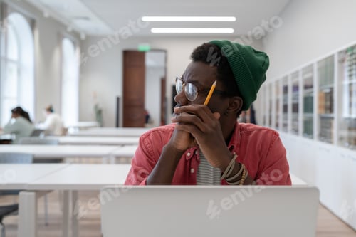 Preview: African student guy distracted from study, sitting at table with laptop in library and looking aside