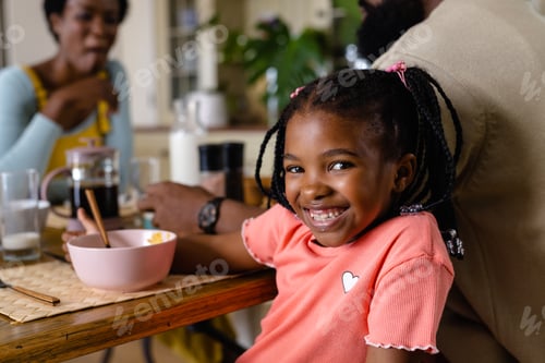 Preview: African american girl smiling at camera while having breakfast with parents at dining table at home