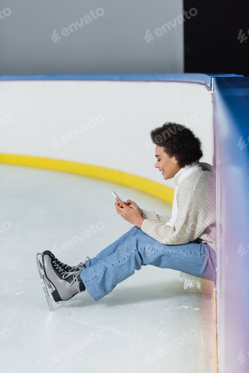 Preview: Side view of cheerful african american woman in ice skates and scarf using smartphone on rink