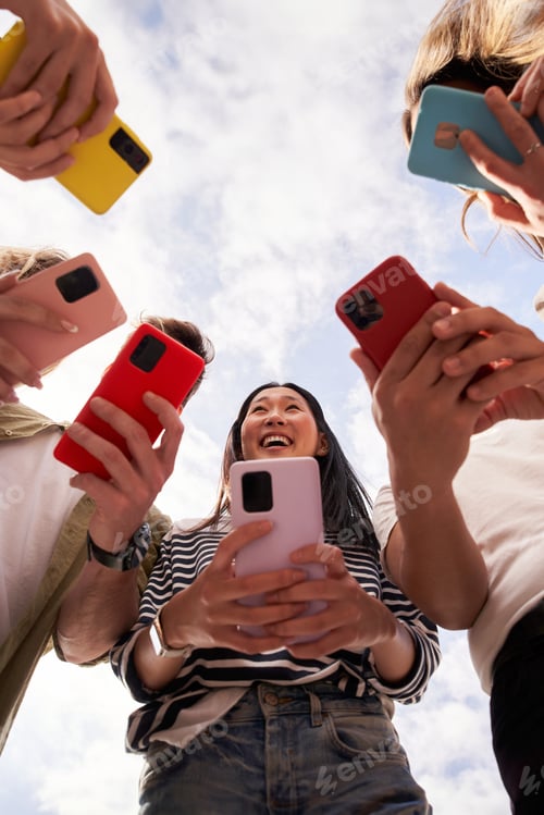Preview: Low angle view of group happy unrecognizable people in circle using colorful mobiles phones outdoor.