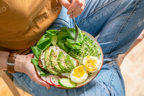 Preview: A woman in jeans sits and holds a plate with fresh food avocado, cucumber, spinach, radish, egg