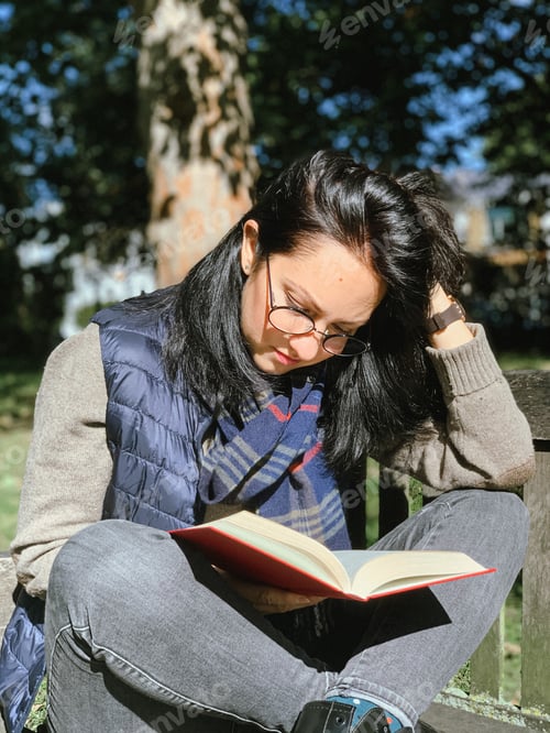 Preview: Young candid woman in casual autumnal clothes siting on a wooden bench in park and reading red book