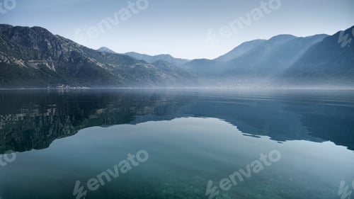 Preview: High mountains reflecting in calm sea waves with rising morning fog or mist