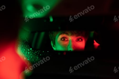 Preview: Eyes of European woman are reflected in automotive mirror riding taxi in evening rainy weather