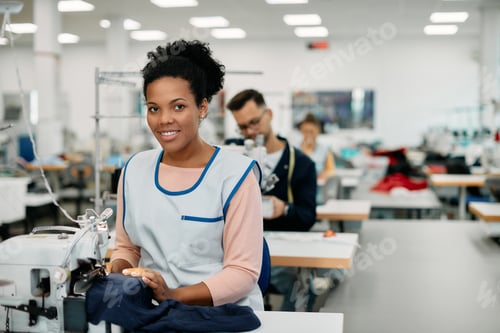 Preview: Happy African American seamstress sewing at clothing design studio and looking at camera.