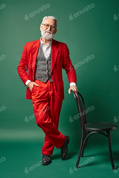 Preview: elegant stylish Santa with beard and glasses standing near chair and hand in pocket, winter concept