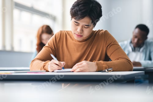 Preview: Asian male student sitting at desk in classroom writing exam