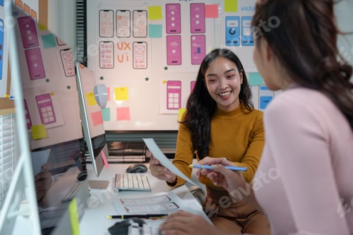 Preview: Two young asian women working as ui/ux designers are discussing mobile app wireframe interface devel
