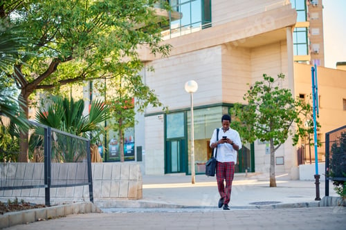 Preview: A Black businessman stands outside a modern skyscraper in an urban area, checking his smartphone