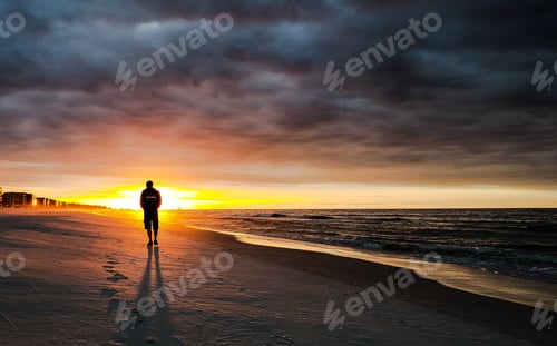 Preview: Solitary silhouetted man on beach at sunrise walking for fitness during golden hour with clouds.