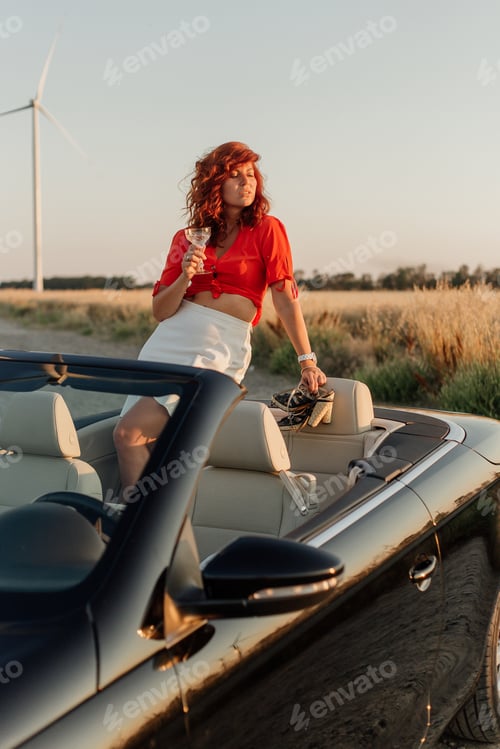 Preview: Beautiful young woman with red hair holding a glass of champagne in a convertible car
