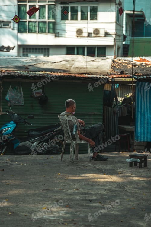 Preview: Vertical shot of a man seated on a grimy plastic chair in a neglected area of Bangkok, Thailand