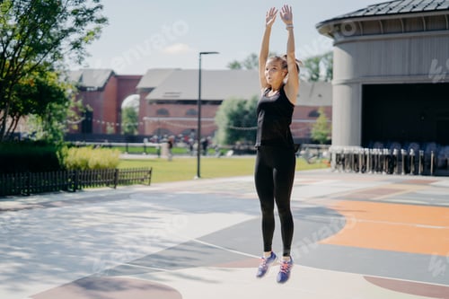 Preview: Active young woman jumps high keeps arms raised up breathes deeply wears black t shirt leggings