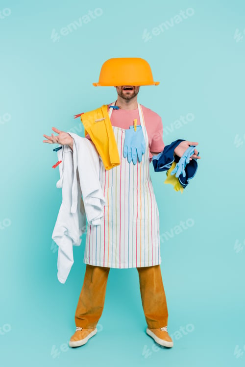 Preview: Shocked unmarried man in clothespins, holding clothes and wash bowl on head on blue background