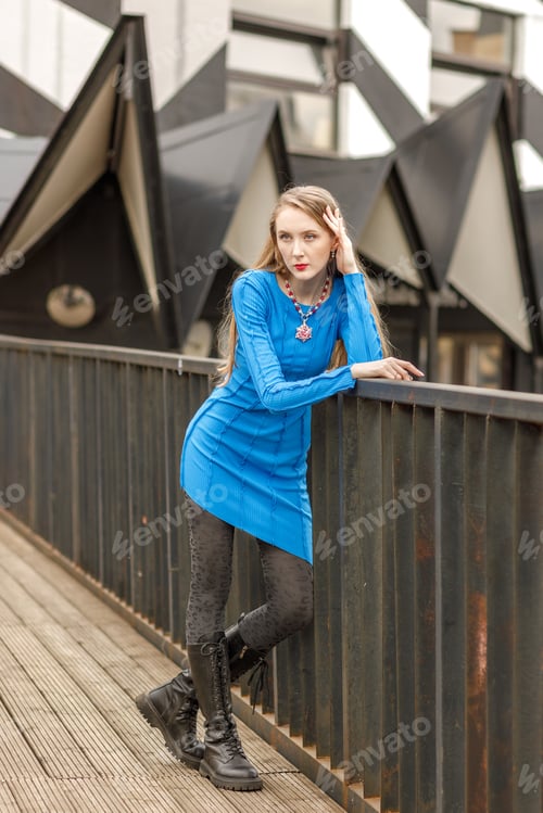 Preview: An elegant girl poses in a blue dress on a bridge between buildings on the street.