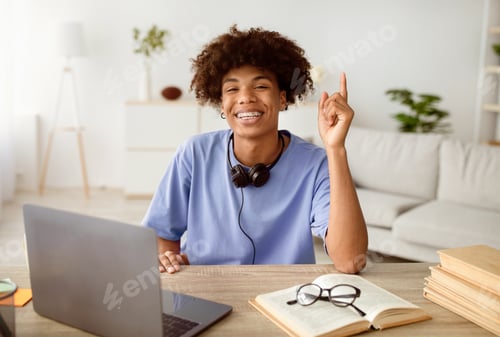 Preview: Happy black teenage student with headphones having creative idea near laptop, gesturing eureka at