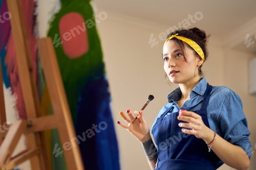 Preview: Portrait of young woman in apron holding paintbrush while working on abstract painting at cozy
