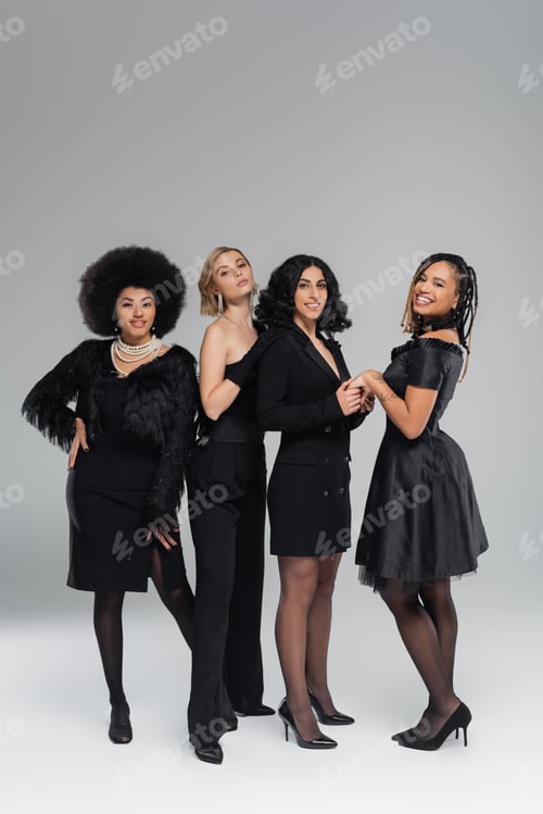 Preview: joyful and elegant multiracial girlfriends in total black clothing smiling at camera on grey