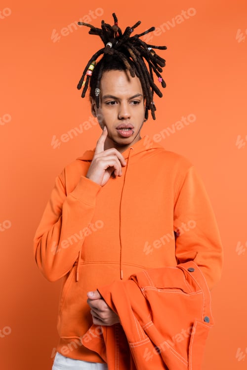 Preview: pensive multiracial man in hoodie holding denim jacket on coral background