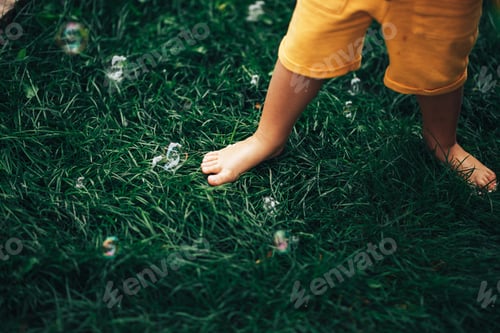 Preview: A small child standing barefoot on a green lawn, a child tramples soap bubbles with his feet