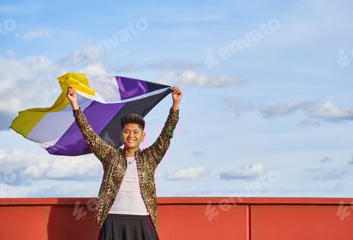 Preview: gender non binary asian person smiling holding gender non binary flag with blue sky background