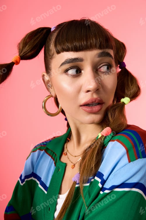Preview: Stylish young woman with playful pigtails and vibrant outfit poses against a pink backdrop