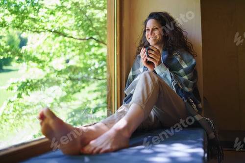 Preview: Happy woman with coffee sitting indoors in a tree house, weekend away and digital detox concept.