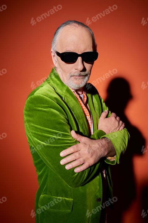 Preview: confident grey haired and bearded senior man standing with folded arms on red and orange background