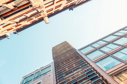 Preview: Upward view of modern high-rise buildings against a clear blue sky with a visible street